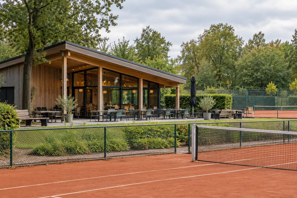 Houten clubhuis met terras naast gravelbanen in een park